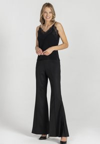 Czarna koszulka na ramiączkach ozdobiona koronką, zestawiona z czarnymi spodniami z wysokim stanem, rozszerzanymi i z plisowanymi detalami. Modelka stoi na neutralnym tle.