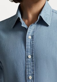 Ljusblå denimskjorta med spetsig krage, som har en knapprad och fem vita knappar. Texturen ser mjuk och något rynkad ut.