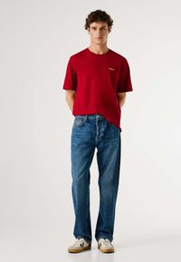 Camiseta roja de manga corta con cuello redondo y logo en el pecho, combinada con jeans azules de pierna recta y zapatillas blancas con detalles en negro.