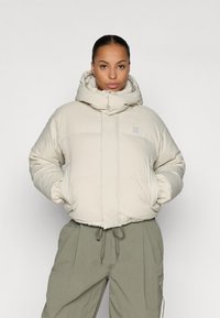 Dik crèmekleurige jas met een hoge kraag, verstelbare boorden en een ritssluiting aan de voorkant. Het heeft horizontale stiksels en een klein logo.