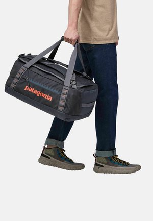Bolsa de deporte gris con el logo naranja de "Patagonia", que cuenta con correas grises, un bolsillo con cremallera y un diseño resistente. Combinada con jeans azules y zapatos coloridos.
