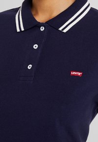 Polo bleu marine avec double rayures blanches sur le col, trois boutons blancs, et petit logo rouge Levi's sur la poitrine.