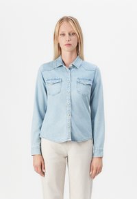 Camisa de mezclilla azul claro con mangas largas, dos bolsillos en el pecho, botón en la parte delantera y dobladillo curvado. Textura suave y diseño clásico de cuello.