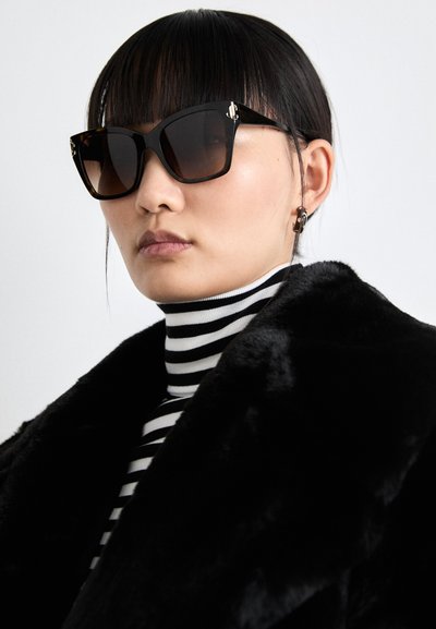 Schwarze übergroße Sonnenbrille mit braunem Verlauf. Begleitet von einem gestreiften Rollkragenpullover und einem schwarzen Kunstfellmantel.