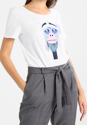 Vrouw draagt een wit T-shirt met kleurrijke abstracte opdruk van een apengezicht, ingestopt in een hoge grijze broek met een strikceintuur.