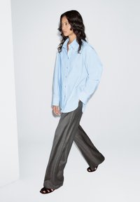 Lichtblauw overhemd met knopen, losvallend model, gecombineerd met grijze wijde broekspijpen. Zwarte sandalen. Minimalistische achtergrond, model lopend.