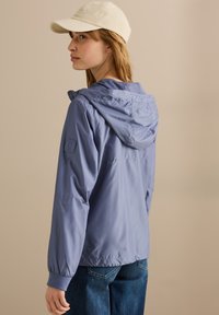 Veste à capuche bleu clair en matériau lisse avec des poignets élastiques. Portée avec une casquette crème et un jean bleu, révélant un design simple.