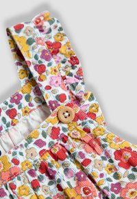 Közelkép virágmintás textil pántokról sárga, piros, rózsaszín virágokkal és zöld levelekkel, amelyek egy kerek fa gombbal vannak rögzítve, fehér háttéren.