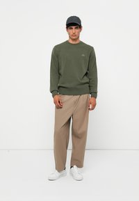 Olive green sweater with ribbed cuffs and hem, paired with beige loose-fit trousers and white sneakers. Black baseball cap completes the look.