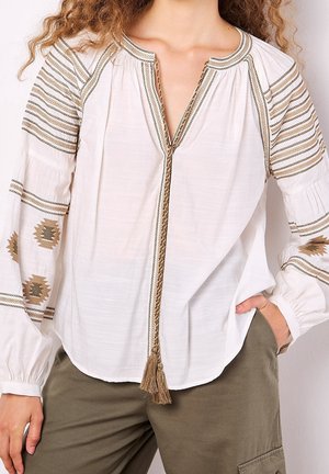 Witte blouse met bruin geborduurde patronen op de mouwen en schouders, kwastje aan de halslijn, gedragen met olijfgroene broek.