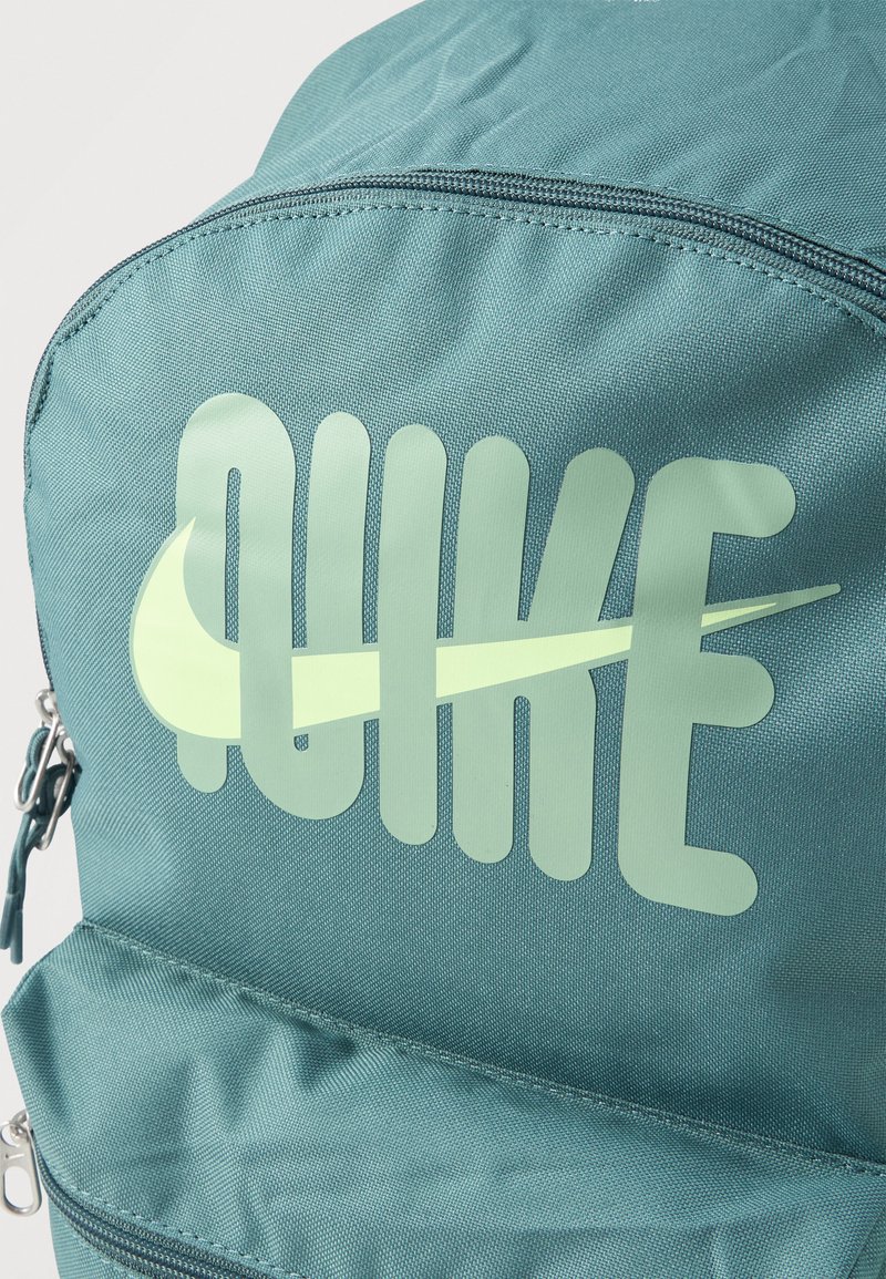 Mochila color verde azulado con texto grande "NIKE" en verde claro y logo swoosh en el bolsillo delantero, cremallera visible y textura de tela.