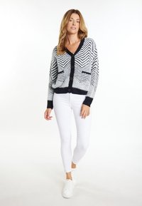 Schwarz-weißer Cardigan mit Zickzack-Muster, V-Ausschnitt, Knopfverschluss und zwei vorderen Taschen, kombiniert mit weißen Leggings und Sneakers.