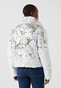 Person facing away wearing a white sequined jacket with silver accents and blue denim jeans against a plain background.