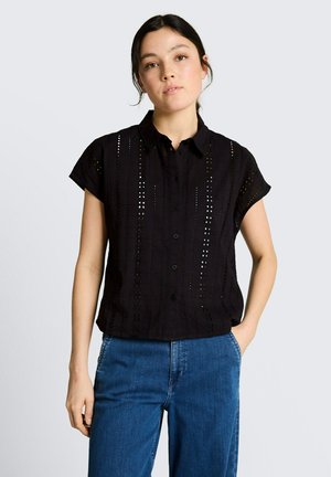 Jeune femme aux cheveux foncés portant un haut noir à manches courtes boutonné avec des détails en œillets et un jean en denim bleu, debout avec une main dans la poche.