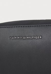 Black textured leather bag featuring embossed "TOMMY HILFIGER" logo in white. Subtle stitching detail along the edges.
