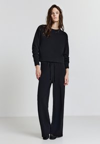 Schwarzer, verkürzter Pullover mit langen Ärmeln, kombiniert mit lockeren schwarzen Hosen. Beide aus einem glatten Material gefertigt und mit einem elastischen Bund ausgestattet.