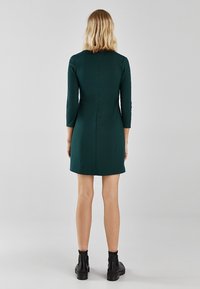 Robe verte ajustée à manches trois-quart, silhouette droite et couture centrale à l'arrière. Portée avec des bottines noires.