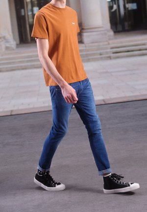 Młoda osoba ubrana w pomarańczowy t-shirt, niebieskie dżinsy i czarne wysokie trampki idąca chodnikiem w pobliżu schodów budynku.