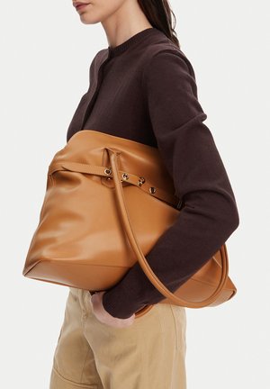 Žena v hnědém svetru a béžových kalhotách drží u boku velkou světle hnědou koženou kabelku s dvojitými uchy.