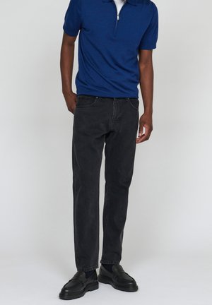 Man wearing a blue short-sleeve zip-up shirt, black jeans, and black leather loafers standing against a gray background.