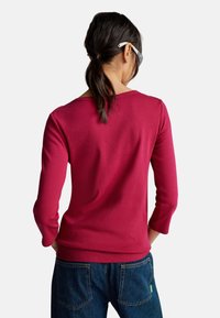 Frau mit dunklen, nach hinten gebundenen Haaren, die ein langärmliges, dunkelrosa Hemd und eine blaue Jeans trägt, von hinten vor einem weißen Hintergrund gezeigt.