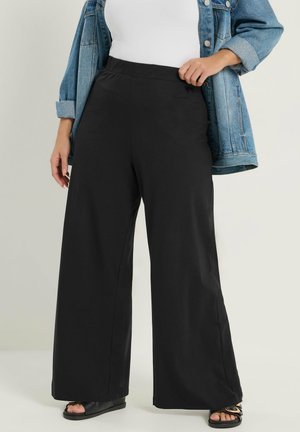 Frau trägt schwarze, weit geschnittene Hose, weißes Oberteil, Jeansjacke und schwarze Sandalen, steht vor einem schlichten Hintergrund.