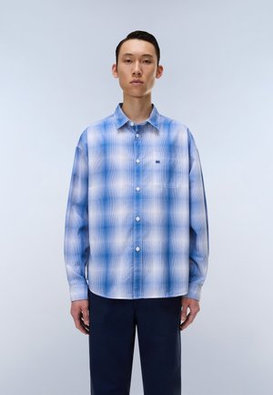 Ung mand iført en blå- og hvidternet skjorte med knapper og mørke bukser, stående mod en ensfarvet lys baggrund.