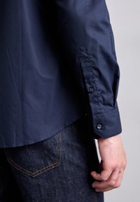 Dunkelblaue Hemd mit glatter Textur, versehen mit einer knöpfbaren Ärmeln und taillierter Passform. Kombiniert mit dunklen Denim-Jeans mit orangefarbener Naht.