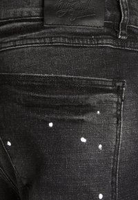 Gros plan sur la poche arrière d'un jean en denim noir avec des éclaboussures de peinture et un patch de marque en cuir au-dessus de la poche.