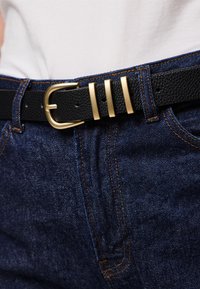 Cinto de couro texturizado preto com uma fivela de metal dourado, apresentando três realces metálicos horizontais; usado sobre um par de jeans de denim azul.