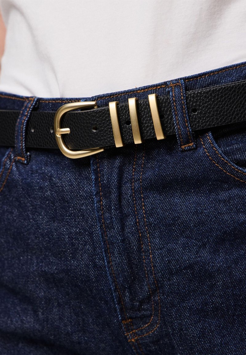 Cinto de couro texturizado preto com uma fivela de metal dourado, apresentando três realces metálicos horizontais; usado sobre um par de jeans de denim azul.