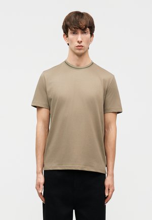 Jeune homme aux cheveux bruns portant un t-shirt beige uni à manches courtes et un pantalon noir, debout devant un fond blanc.