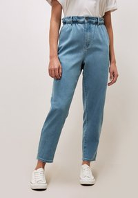 Pantaloni in denim blu chiaro con vita elastica e vestibilità rilassata, dotati di chiusura frontale con bottone e tasche laterali. Indossati con sneakers bianche.