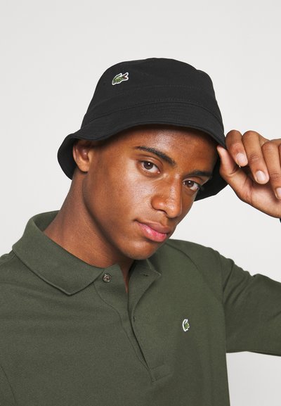Chapeau seau noir avec un logo Lacoste vert, en tissu structuré, texture douce et un large bord arrondi. Porté avec un polo vert foncé.