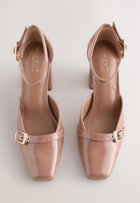 Scarpe con tacco a blocco in vernice color nude, punta quadrata, cinturino alla caviglia e dettaglio fibbia color oro. Finitura liscia e design minimalista.