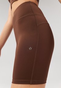 Brown athletic shorts made of stretchy material, featuring a high waist, flatlock seams, and a logo on the lower side.
