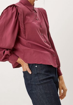 Person iført en langærmet bærfarvet skjorte med knapper og puffede ærmer samt mørkeblå denimjeans, hånd i lommen.