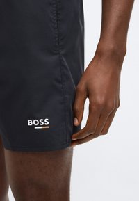Čierne plavecké šortky s hladkou textúrou, s bielym logom "BOSS" a farebnými akcentovými pásmi na ľavej strane. Detailný záber na držiacu ruku.