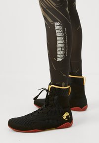 Black and red high-top sports shoes with yellow accents worn with black leggings featuring "Conquer Your Future" text and abstract design.