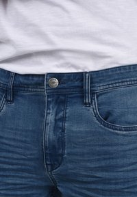 Tamno plavi traperice od denima s glatkom teksturom, s metalnim zatvorenjem na dugme i tradicionalnim dizajnom s pet džepova.