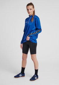 Maglietta azzurra a maniche lunghe con mezza zip e motivo a zig zag nero sulle maniche, abbinata a pantaloni corti neri aderenti e scarpe da ginnastica blu.
