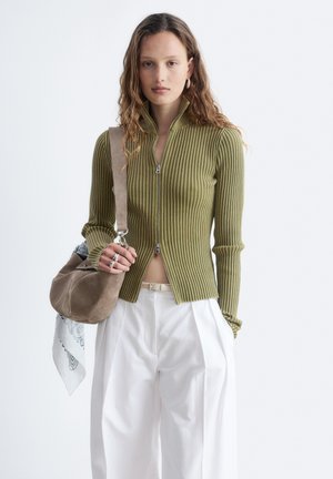 Jeune femme portant un pull côtelé zippé vert olive et un pantalon blanc taille haute, portant un sac à bandoulière en daim beige avec une écharpe blanche.