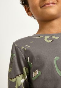 Grå bomullsskjorta med gröna dinosaurieavtryck, inklusive T. rex och brontosaurus, med palmträdsmotiv och en mjuk textur.