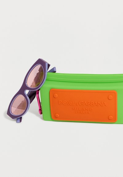 Μωβ γυαλιά ηλίου cat-eye ακουμπισμένα σε μια πράσινη και πορτοκαλί θήκη με το λογότυπο Dolce & Gabbana.