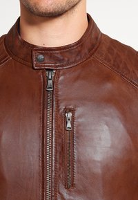 Brun lädermotorcykeljacka med hög krage, exponerad framdragkedja och en liten ficka med dragkedja på bröstet. Har texturerad sömnad.