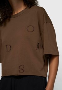 Persona vistiendo una camiseta corta marrón y holgada con letras bordadas D, O, A, S y pantalones negros, sobre un fondo claro y liso.