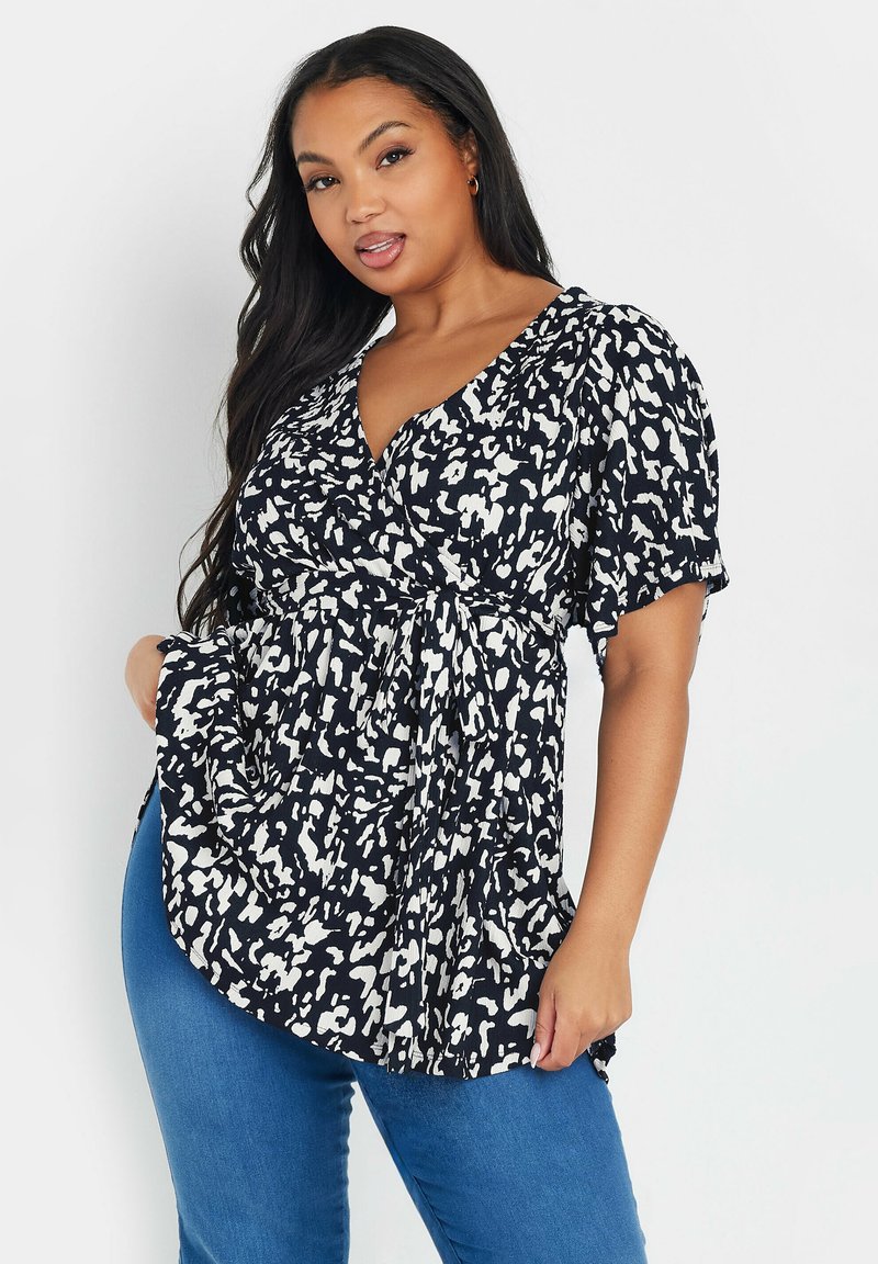 Top blu scuro con motivi astratti bianchi, maniche a sbuffo corte, scollatura a V e dettaglio in vita annodato, abbinato a jeans blu.