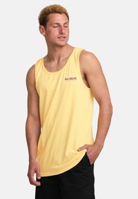 Camiseta sin mangas amarilla con escote redondo, hecha de algodón suave, con un pequeño logo burdeos en el pecho, combinada con pantalones cortos negros.