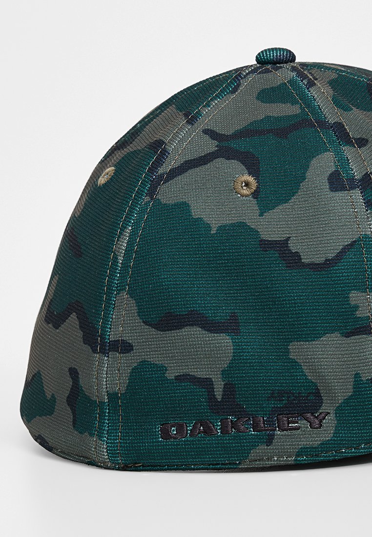 Gorra con patrón de camuflaje en verde y gris, fabricada con un tejido texturizado. Presenta un logo bordado y un visera curvada. Detalle de ventilación metálica visible.