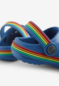 Modre gumijaste cokle imajo dvignjen podplat, mavrično črtasto trak po zgornjem delu in okroglo blagovno znamko na strani.